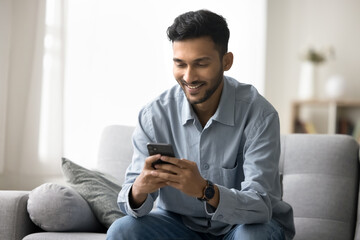 Good news in message. Smiling confident millennial man relax at living room sit on couch use cellphone device to send get sms chat in messenger enjoy virtual dating in web app check social media pages