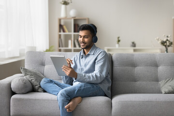 Interactive communication. Smiling confident young male sit on sofa at living room in modern digital headset speak by video call using tablet study online from home chat in conference app. Copy space