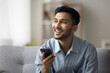 © fizkes - Smiling young man relax at living room sit on comfy couch hold mobile phone think about good news received in email dictate voicemail message speak audio command to virtual ai assistant. Copy space