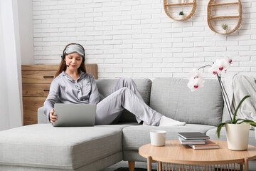 Poster - Young woman in pajamas and headset working with laptop on sofa at home. Remote work concept