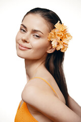 Wall Mural - Young woman with a flower in her hair, wearing an orange dress, smiling gently, captured in a soft light background, conveying a fresh summer vibe.