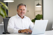 © Azfaribn - Senior businessman working on a laptop in a simple office setting, wearing a white shirt, seated at a plain desk with minimal decor