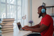 © Tj - Focused black student wearing headphones, sitting at a desk and typing on a laptop, studying online with books nearby, showcasing modern education and self-improvement