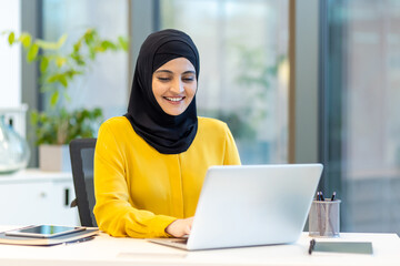 Wall Mural - Successful businesswoman in hijab working with laptop inside office at workplace. Arab woman smiling happily typing on keyboard.