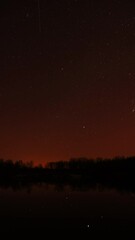 Wall Mural - Real Colorful Night Stars Above Lake River. Natural Starry Sky Background Backdrop. Time Lapse. Vertical Shot. Glowing Stars Reflected In Lake River Water. Natural Starry Sky Background Backdrop