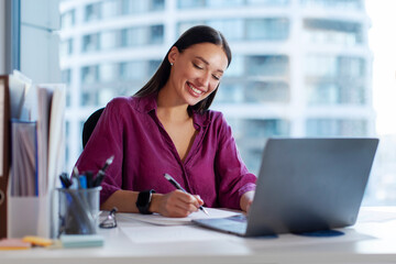 Happy European business woman CEO working on laptop and taking notes, sitting at desk in office. Female professional manager using pc digital computer