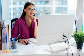 Happy female business professional working at workstation monitor, sitting at desk and smiling, looking at screen, office interior