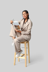 Wall Mural - Beautiful young African-American businesswoman with dreadlocks holding magazine and sitting on stool against grey background