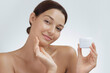 © Buyanskyy Production - A young woman with flawless skin smiling while holding a skincare cream jar and gently touching her face. Perfect for beauty, skincare, cosmetics, and self-care promotional content.