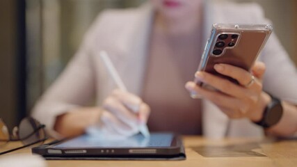A woman is sitting at a table with a tablet and a cell phone in front of her. She is using the cell phone to text while the tablet is open