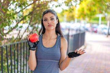 Wall Mural - Young pretty sport woman with an apple at outdoors making doubts gesture while lifting the shoulders