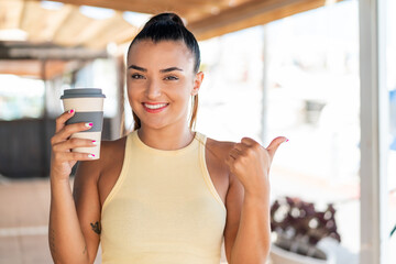 Wall Mural - Young pretty woman holding a take away coffee at outdoors pointing to the side to present a product