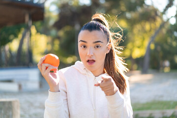 Poster - Young pretty brunette woman holding an orange at outdoors surprised and pointing front