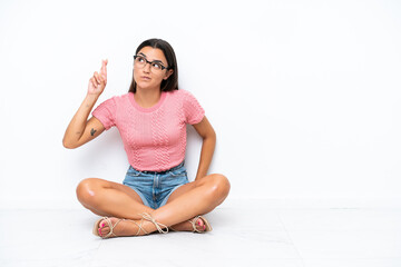 Wall Mural - Young caucasian woman sitting on the floor isolated on white background with fingers crossing and wishing the best