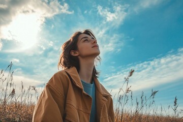 Stylish woman breathing fresh air, enjoying nature in a field with dry grass and looking up at beautiful cloudy sky