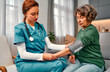 © HBS - Young female doctor in scrubs with stethoscope around neck measuring blood pressure of elderly woman at home while she is sitting on sofa. Doctor's visit to home. Checking blood pressure.