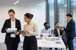 © Liubomir - Two businesswomen standing inside the office at their workplace, office workers inside the meeting room discussing documents and financial figures, in business suits.