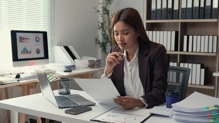 Wall Mural - Young Asian businesswoman reviewing paperwork and using laptop at workplace, demonstrating focus and professionalism in corporate environment