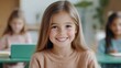 © Y_Malashkevych - A joyful girl around eight years old is smiling broadly at the camera while sitting at a green desk in a classroom. The soft blurred background features other children engaged in activities