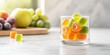 © Y_Malashkevych - A small glass container holds colorful fruit-shaped multivitamin gummies on a light stone kitchen surface with blurred grapes and apples in the background enhancing the vibrant colors