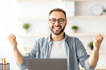 Young guy making YES gesture in front of laptop at home office. Millennial man reaching success, working with portable pc, making profitable deal, signing online contract, celebrating achievement