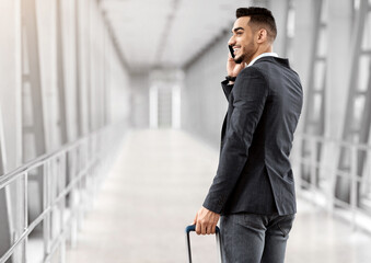 Wall Mural - After Arrival. Smiling Arab Businessman Talking On Cellphone While Passing Airport Gate, Cheerful Middle Eastern Man Walking With Suitcase In Terminal And Enjoying Mobile Call, Rear View