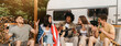 © Prostock-studio - Cheerful interracial friends playing guitar and singing, holding American flag, drinking beer, sitting near RV, celebrating Independence Day, having fun camping trip on summer vacation