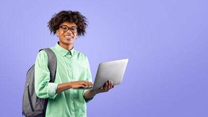 Online education concept. Happy black student guy with backpack and laptop computer, standing over blue background and smiling at camera. Online homework and e-learning