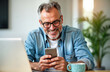 © Maryna - Smiling middle-aged man checks news on phone at home. Happy mature adult in casual attire sits at home office desk. Uses smartphone to read news. Confident older man relaxed, happy. Home office