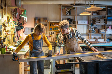 Carpentry colleagues handle circular saw station, measuring wooden beam with precision. Collaborative effort, female mentor teaching newby, shared goals, handcrafted wooden goods production for shop