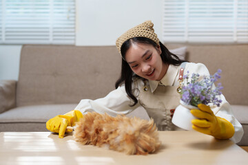 Wall Mural - Young smiling housekeeper wearing yellow gloves is cleaning a coffee table with a feather duster and holding a decorative plant in the living room