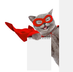 Aufkleber - Happy cat wearing superhero costume shows blank notepad in it paw and looks from behind blank white banner. Isolated on white background