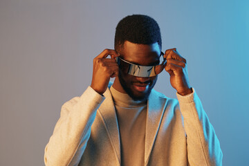 Stylish man wearing futuristic sunglasses in a modern outfit with a confident smile against a blue and gray background, ideal for fashion and technology themes