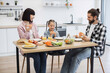 © sofiko14 - Caucasian family with young girl having breakfast with pizza. Parents and child sitting at table enjoying meal. Bright kitchen setting with fruits and vegetables.