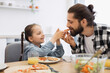 © sofiko14 - Caucasian father and daughter sharing pizza breakfast. Smiling girl with braided hair feeds bearded dad. Bright kitchen atmosphere with natural light, orange juice, and salad on table.