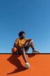 © VISTA by Westend61 - Afro man with basket ball sitting on orange wall under blue sky