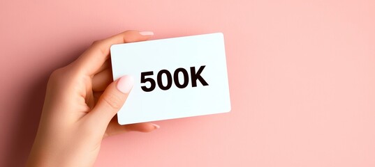 Female Hand Holding a White Card With 500k Written On It, Isolated on a Pink Background