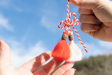 Martisor or Martenitsa in female hands on blue sky background, copy space. Bulgarian, Romanian and Moldovan tradition giving trinkets made of white and red threads, beginning of spring, first of March