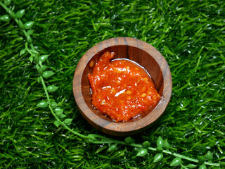 Wall Mural - Indonesian traditional chili sauce named sambal terasi. crushed red chili peppers and mixed with shrimp paste, garlic, onion, and spices, served in a small wooden bowl on green synthetic grass base.