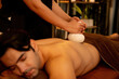 © InfiniteFlow - Hot herbal ball spa massage body treatment, masseur gently compresses herb bag on man body. Tranquil and serenity of aromatherapy recreation in warm lighting of candles at spa salon. Quiescent