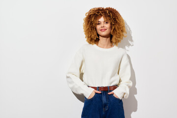 Wall Mural - Smiling young woman with curly hair wearing a cozy white sweater and high waisted jeans against a minimalistic white background, exuding confidence and warmth