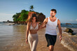 © artiemedvedev - Fit couple hold hands, walk on tropical beach. Smiling woman in bikini, man in tank top enjoy seaside. Freelancers unwind on sandy shore, balance work, travel lifestyle. Remote job, leisure together.