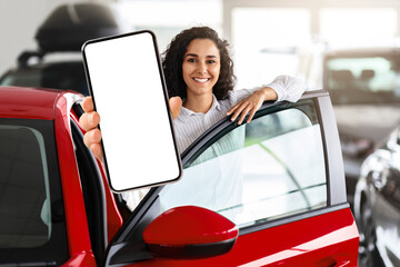 Positive millennial middle eastern woman standing by brand new yellow car, showing smartphone with empty screen and smiling at camera, mockup, renting auto on Internet. Car dealership online