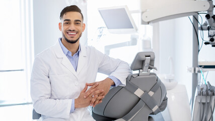 Wall Mural - Dental Services. Portrait Of Professional Arab Stomatologist Doctor Posing At Workplace In Modern Clinic Interior, Friendly Middle Eastern Dentist In Uniform Smiling At Camera, Ready For Check Up