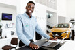 © Prostock-studio - Portrait of happy car salesman standing at work desk, smiling at camera, using laptop and touch pad in auto dealership. Positive black automobile manager posing at workplace in showroom store