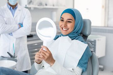 Wall Mural - Portrait Of Happy Muslim Woman Patient Looking At Mirror After Treatment In Dental Clinic, Smiling Islamic Lady In Hijab Sitting In Chair In Stomatological Cabinet And Enjoying Her Perfect Teeth