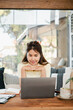 © Satori Studio - Young woman using a laptop in a cozy cafe, concentrating on work, with notebooks and coffee on the table.