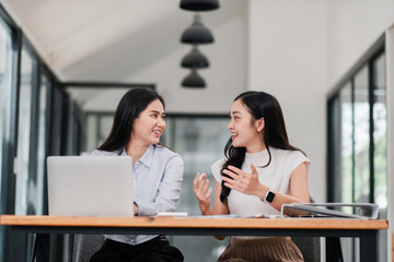 Two businesswomen having a conversation at a desk in a contemporary office setting, showcasing teamwork and collaboration.