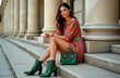 © Pete - Stylish woman sits on urban steps. Wears colorful spring summer dress. Elegant outfit includes trendy boots, matching green handbag. Fashionable model poses in city. Outdoor photoshoot. High fashion