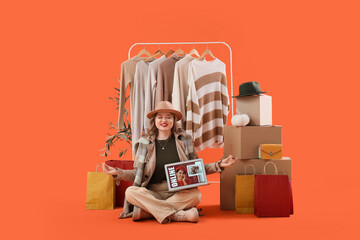 Wall Mural - Beautiful young woman in autumn clothes with laptop, shopping bags and cardboard boxes meditating on orange background. Online shopping concept